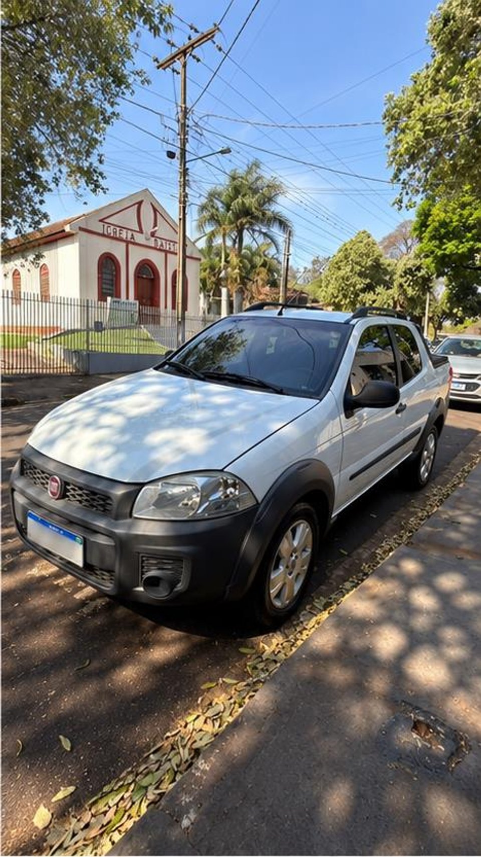 Fiat Strada branca anunciada vista de frente em três quartos, estacionada em rua arborizada